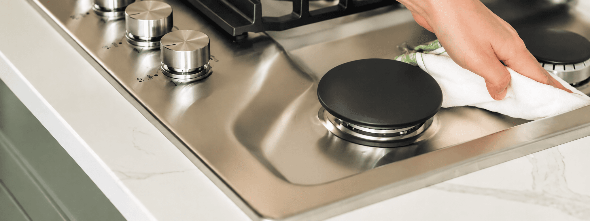 Hand wiping a stainless steel gas stovetop with a white cloth near a black burner.